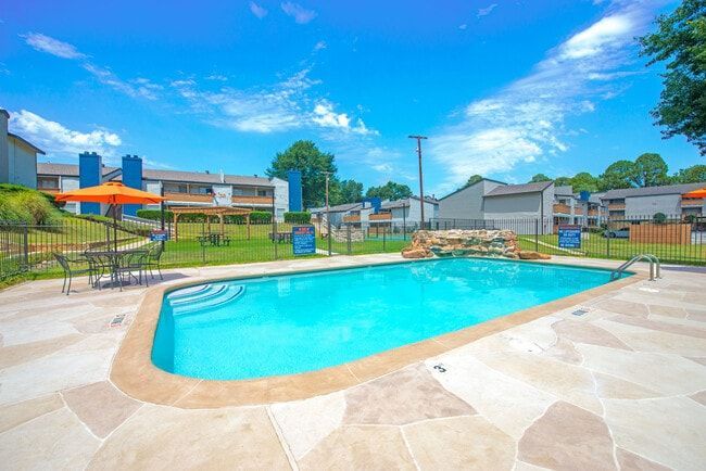 Swimming pool with blue water, surrounded by stone paving and a fence. Apartment buildings in the background.