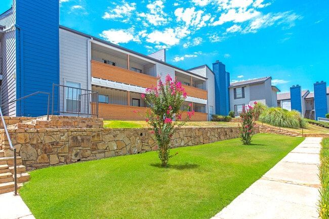 Apartment complex with blue accents, green grass, stone wall, and bright blue sky.