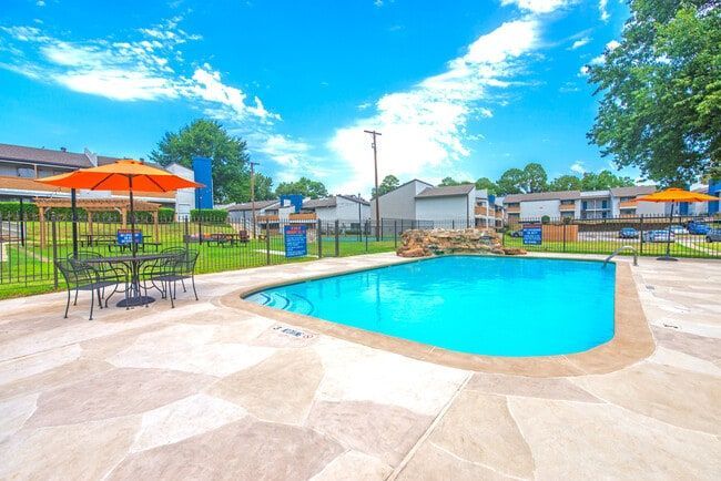 Pool area with blue water, orange umbrellas, table, chairs, and buildings under a blue sky.