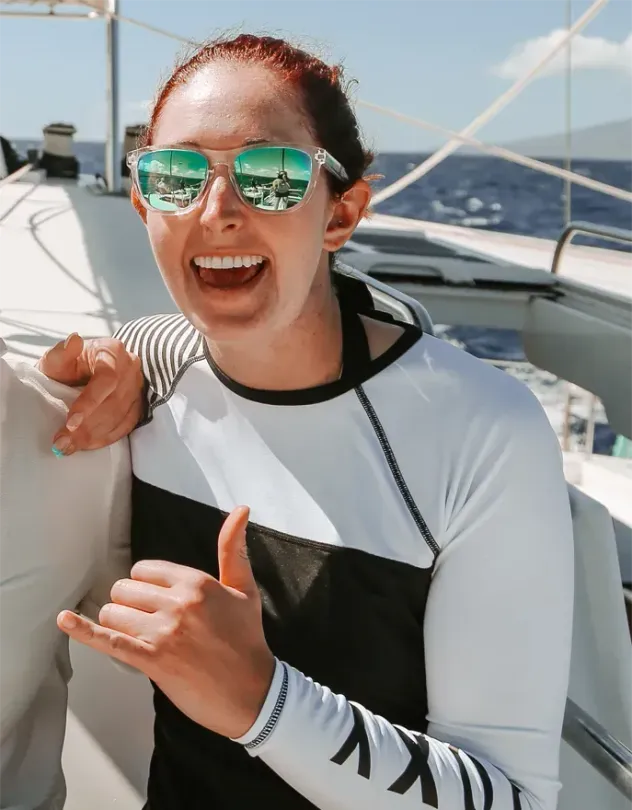 Smiling person in sunglasses on a sailboat, wearing a white and black top, with mountains in the background