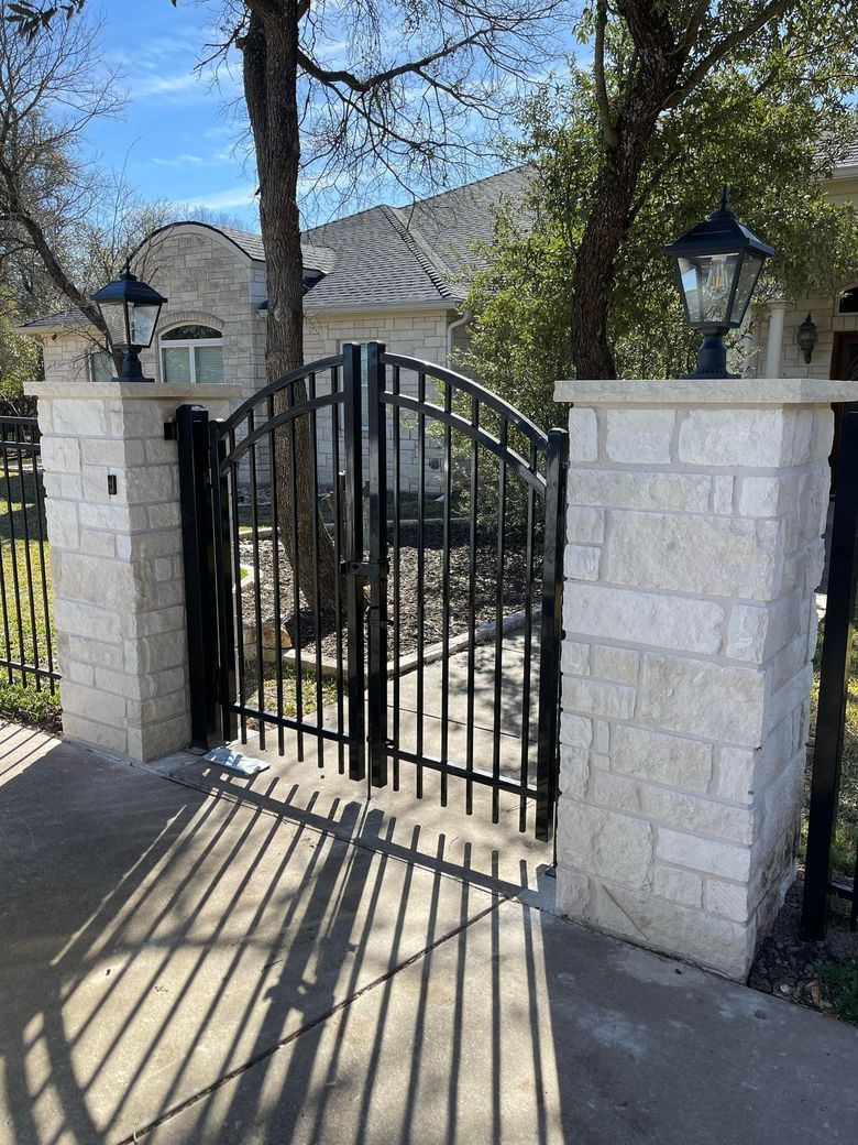 Black wrought-iron gate between white stone pillars beside a driveway, with house and trees behind.