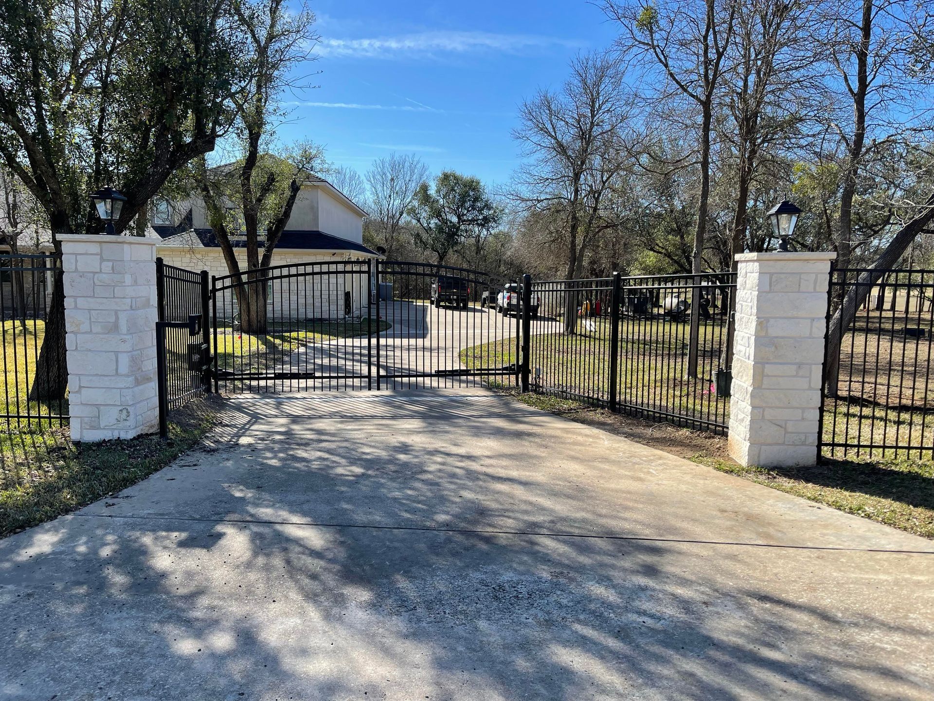 Black metal gate between white stone pillars at a driveway entrance, with trees and a house behind.