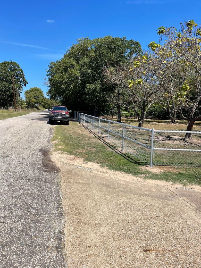 Rural dirt road with parked car, fenced yard, and trees under a clear blue sky