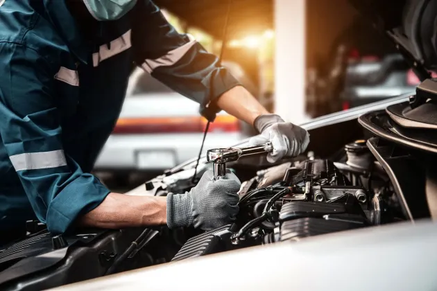 Mechanic in blue coveralls inspecting a car engine in a garage, wearing gloves under bright shop lights
