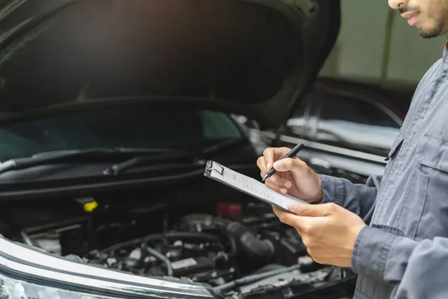 Mechanic writing on a clipboard beside an open car hood in a garage