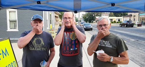 Three men are standing under a tent on the sidewalk covering their mouths.