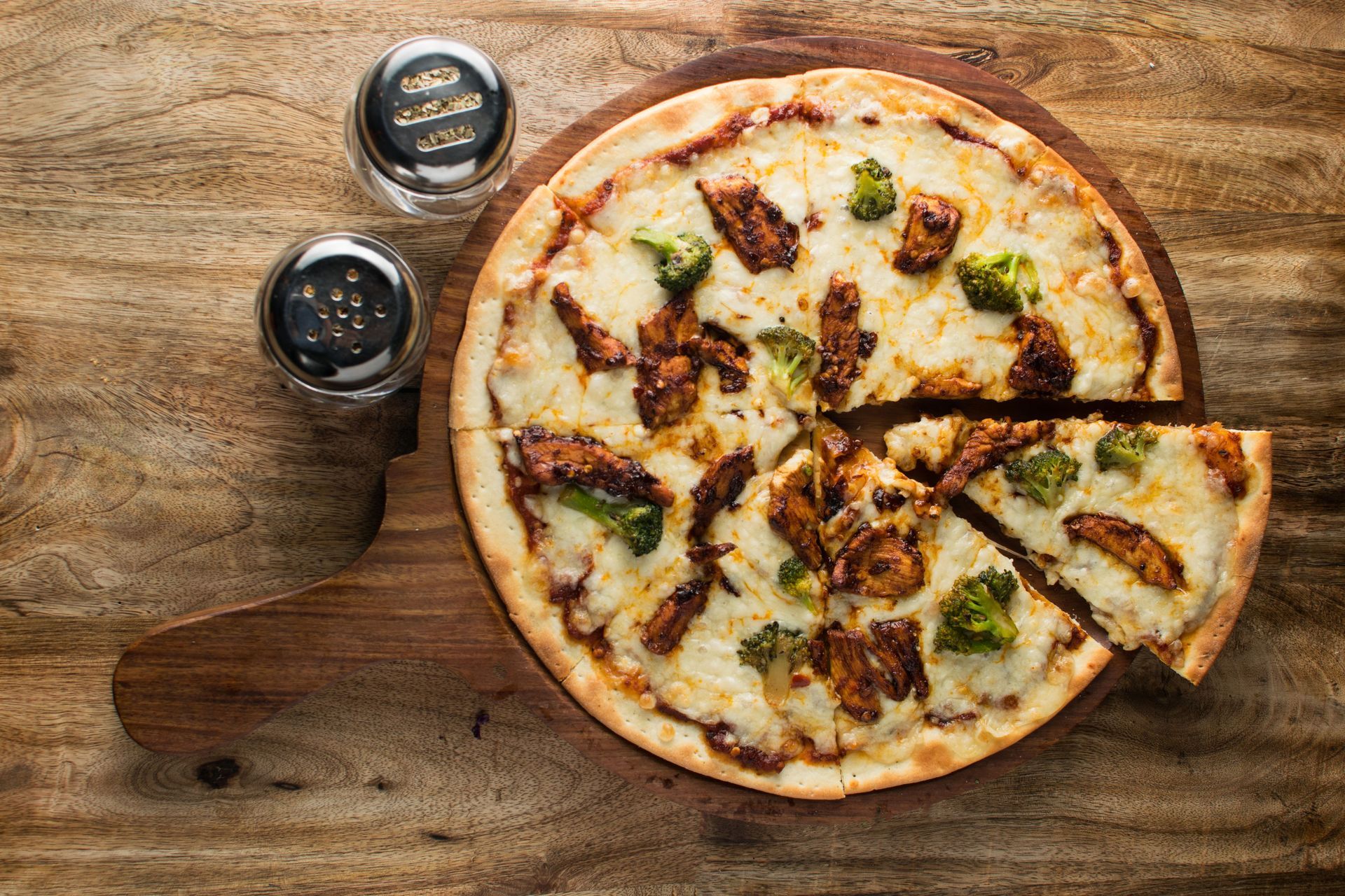 Two pizzas on a wooden table, one slice removed, with two small condiment jars nearby