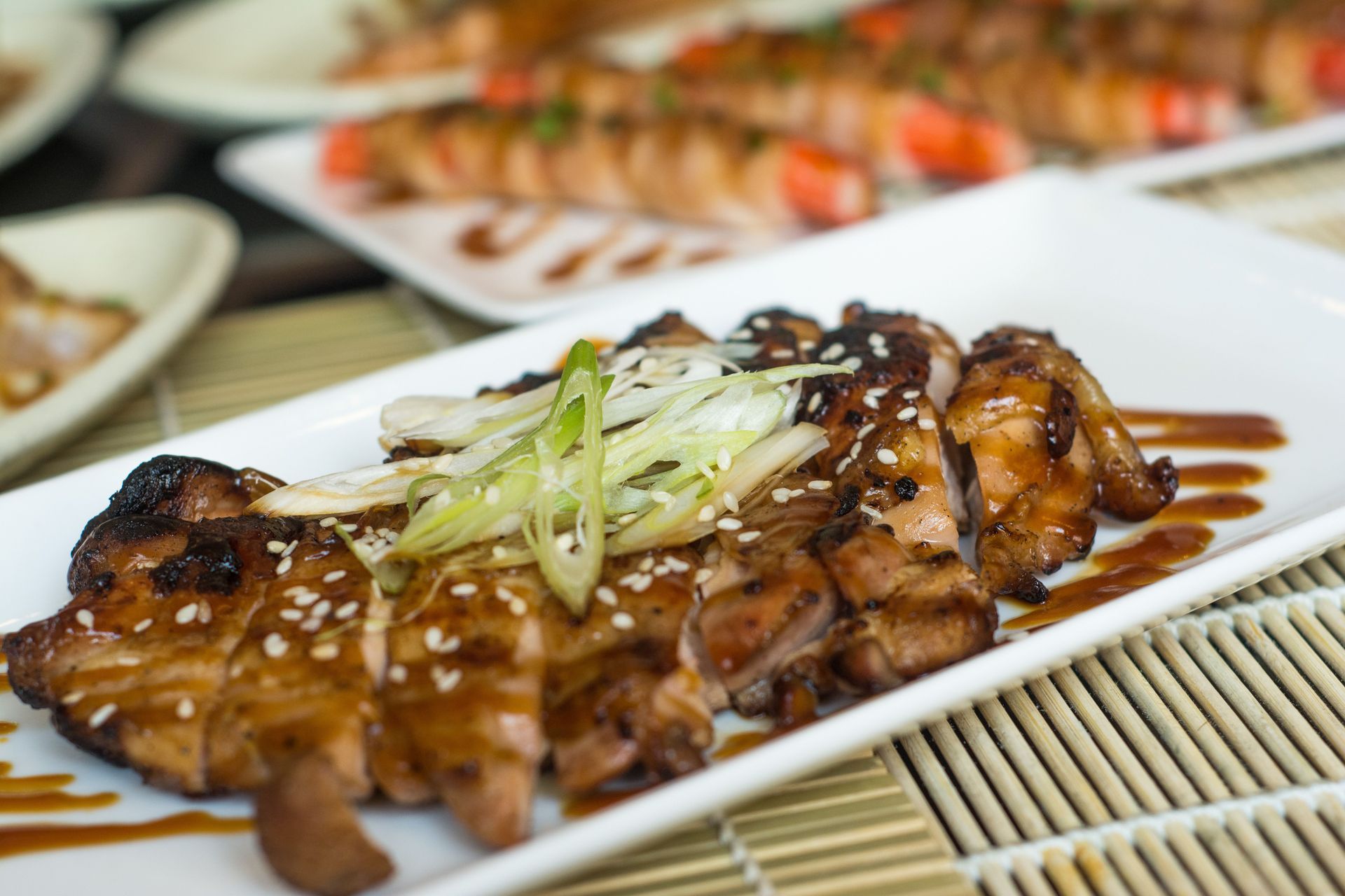 Glazed grilled chicken skewers topped with scallions on a bamboo mat, with sushi plates blurred in the background