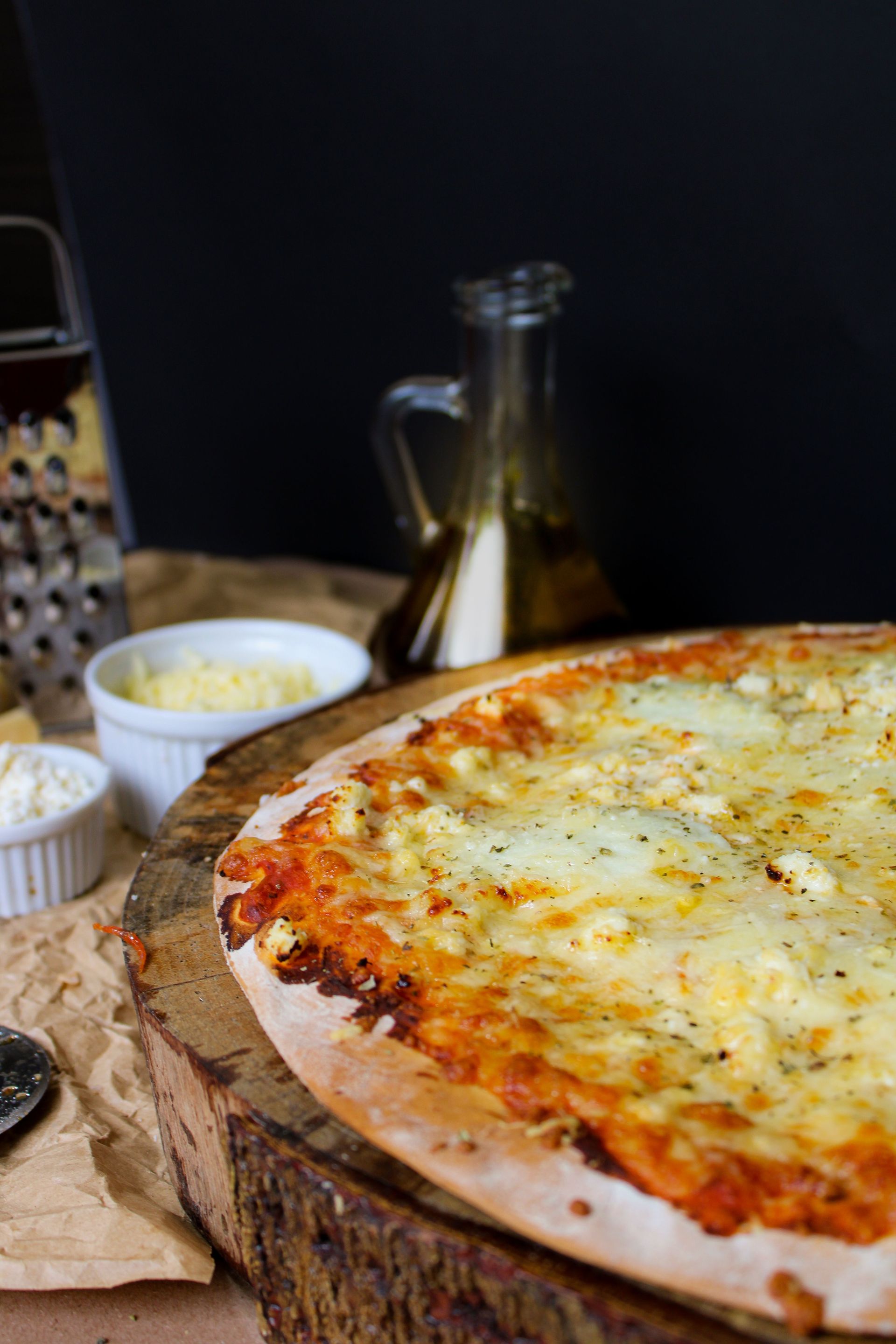 Cheese pizza on a wooden board with small bowls of toppings and a bottle in the background