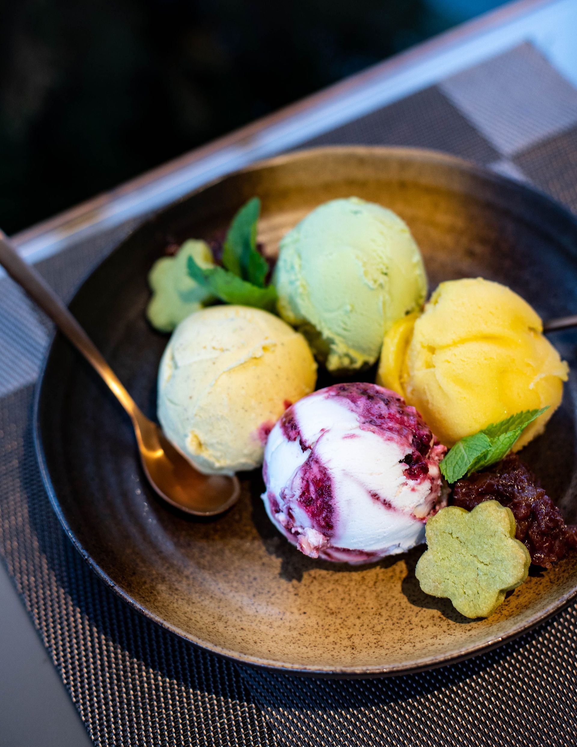 Bowl of four colorful scoops of ice cream with a spoon on a woven placemat.