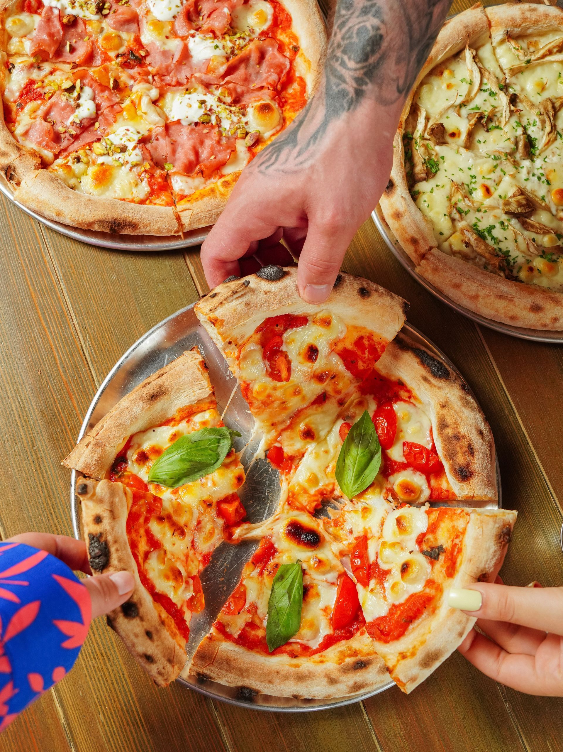 Hands reaching for slices of pizza on a table with three pies, including one with basil and tomato sauce.