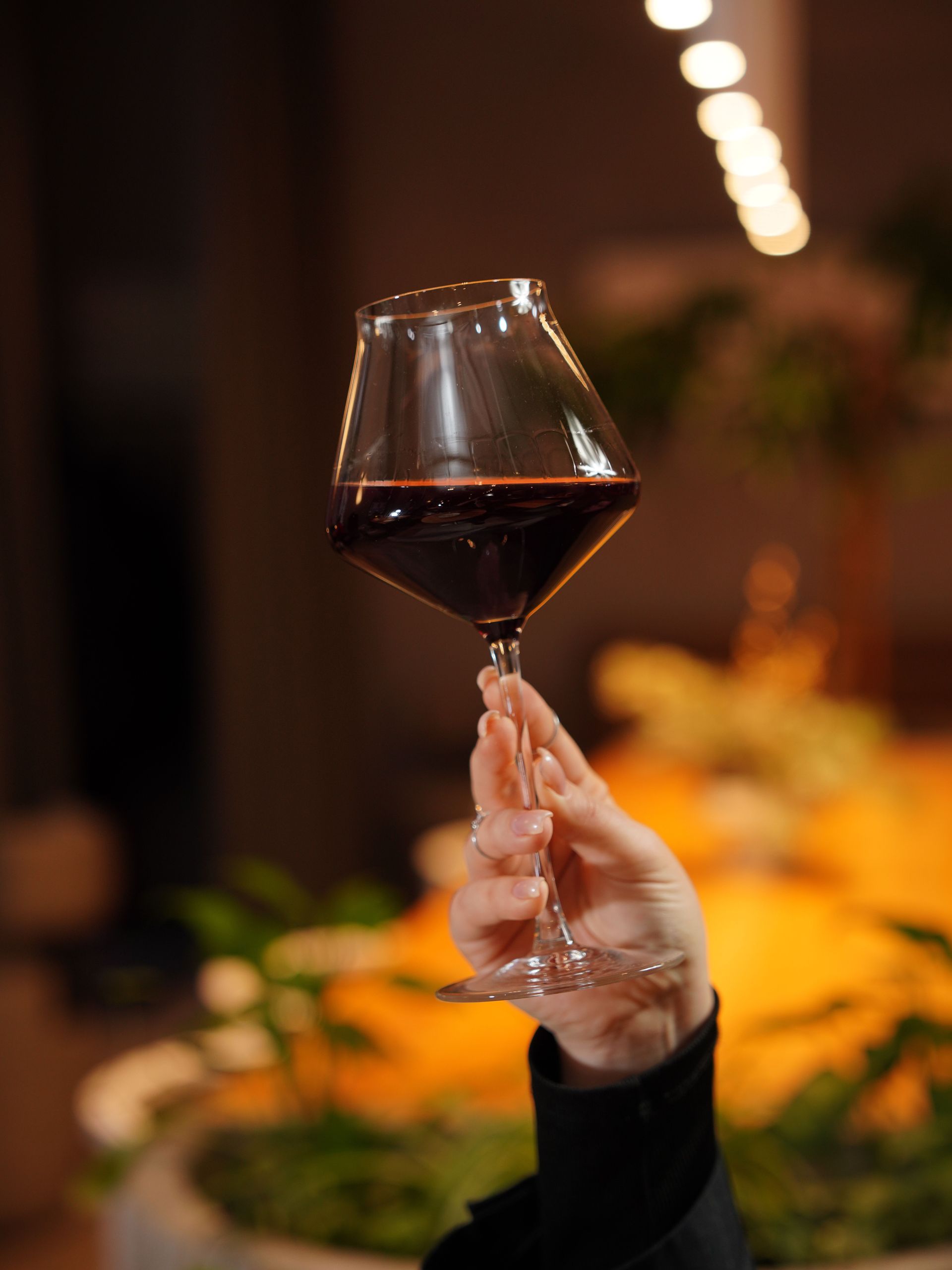 Hand holding a stemmed wine glass with red wine at a warmly lit dinner table