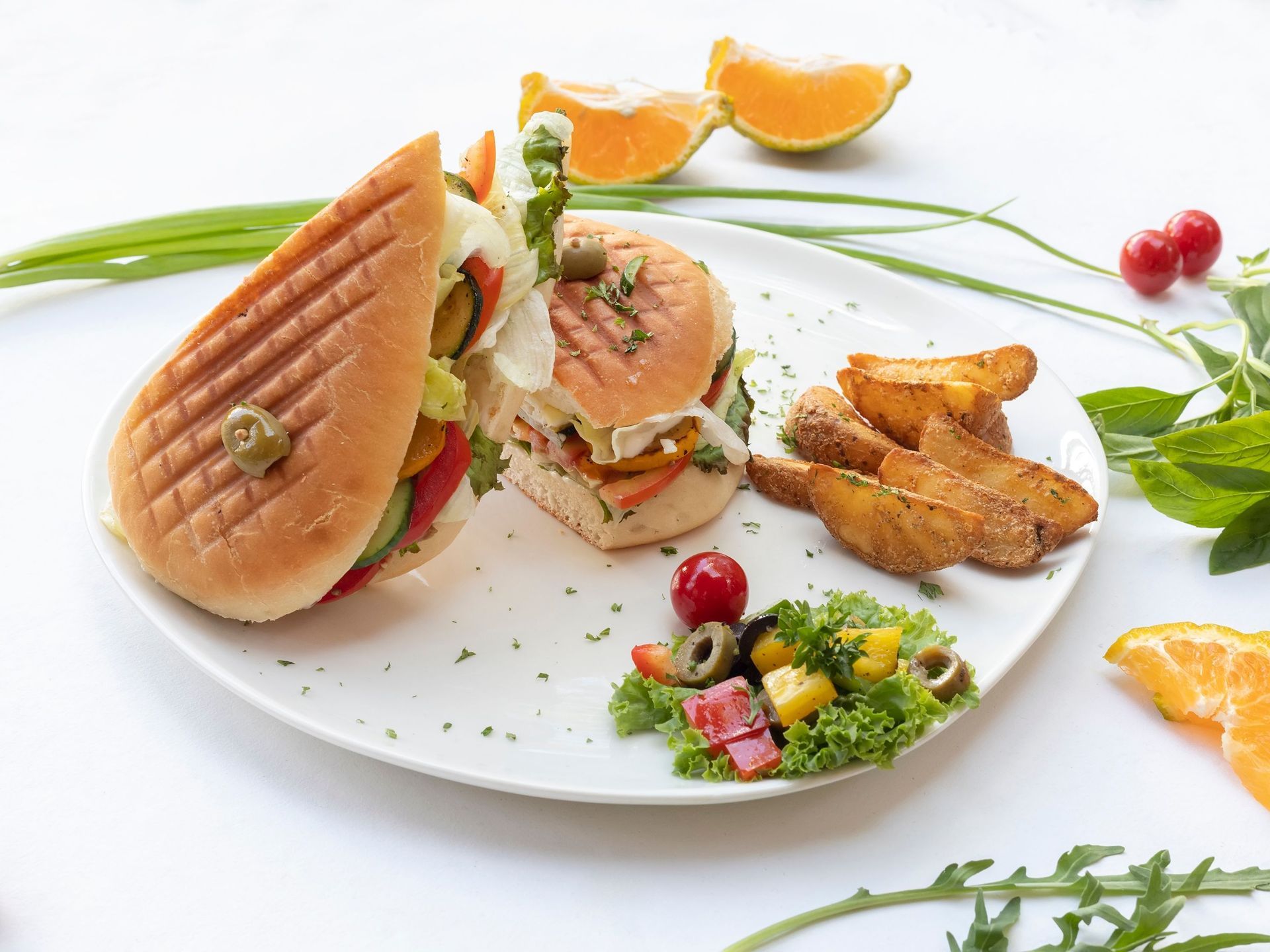 Plated gourmet salmon sandwich with vegetables, potato wedges, and orange slices on a white background