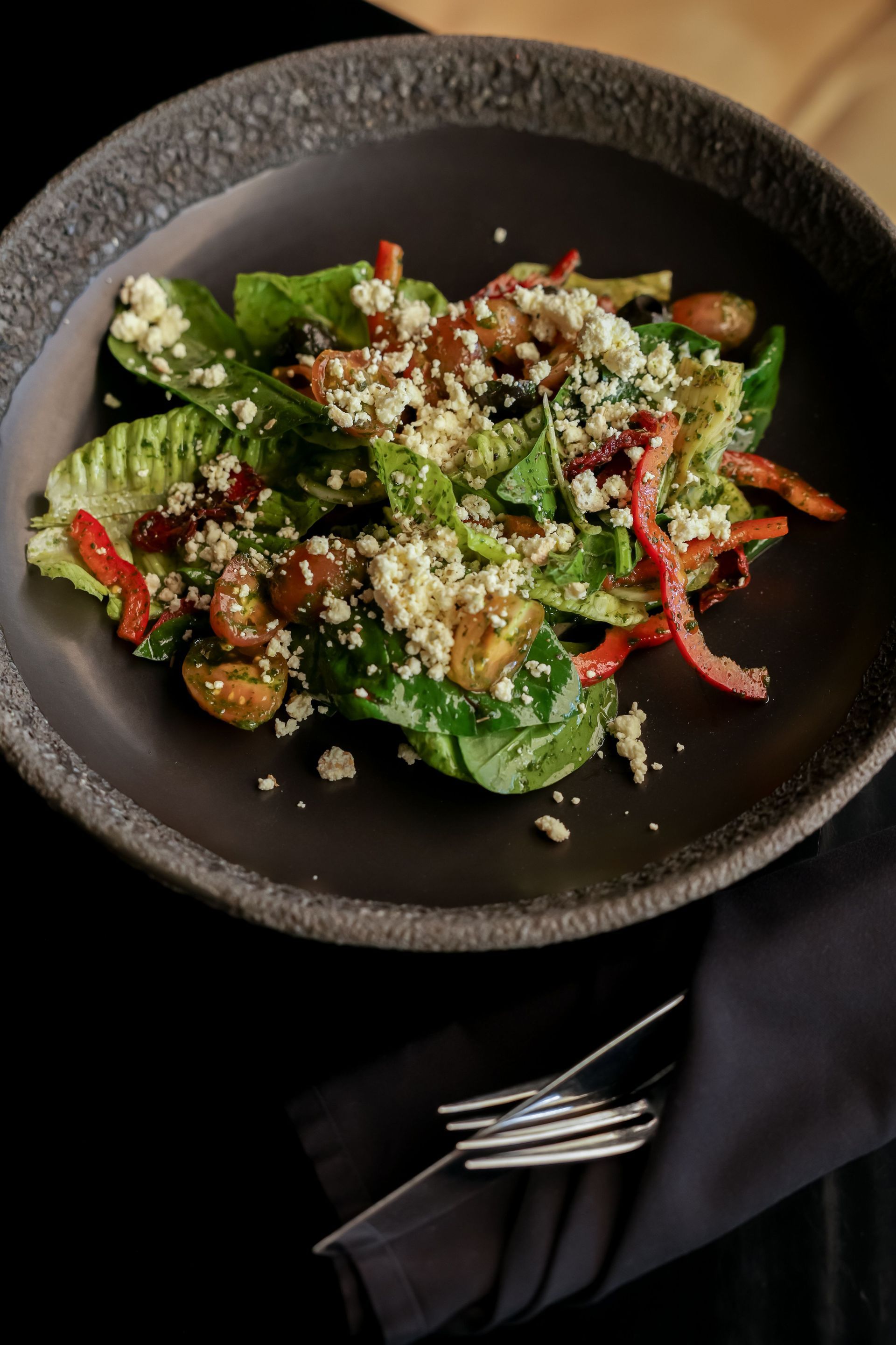 Black bowl of salad with greens, red peppers, and grated cheese on a dark table with a fork and napkin