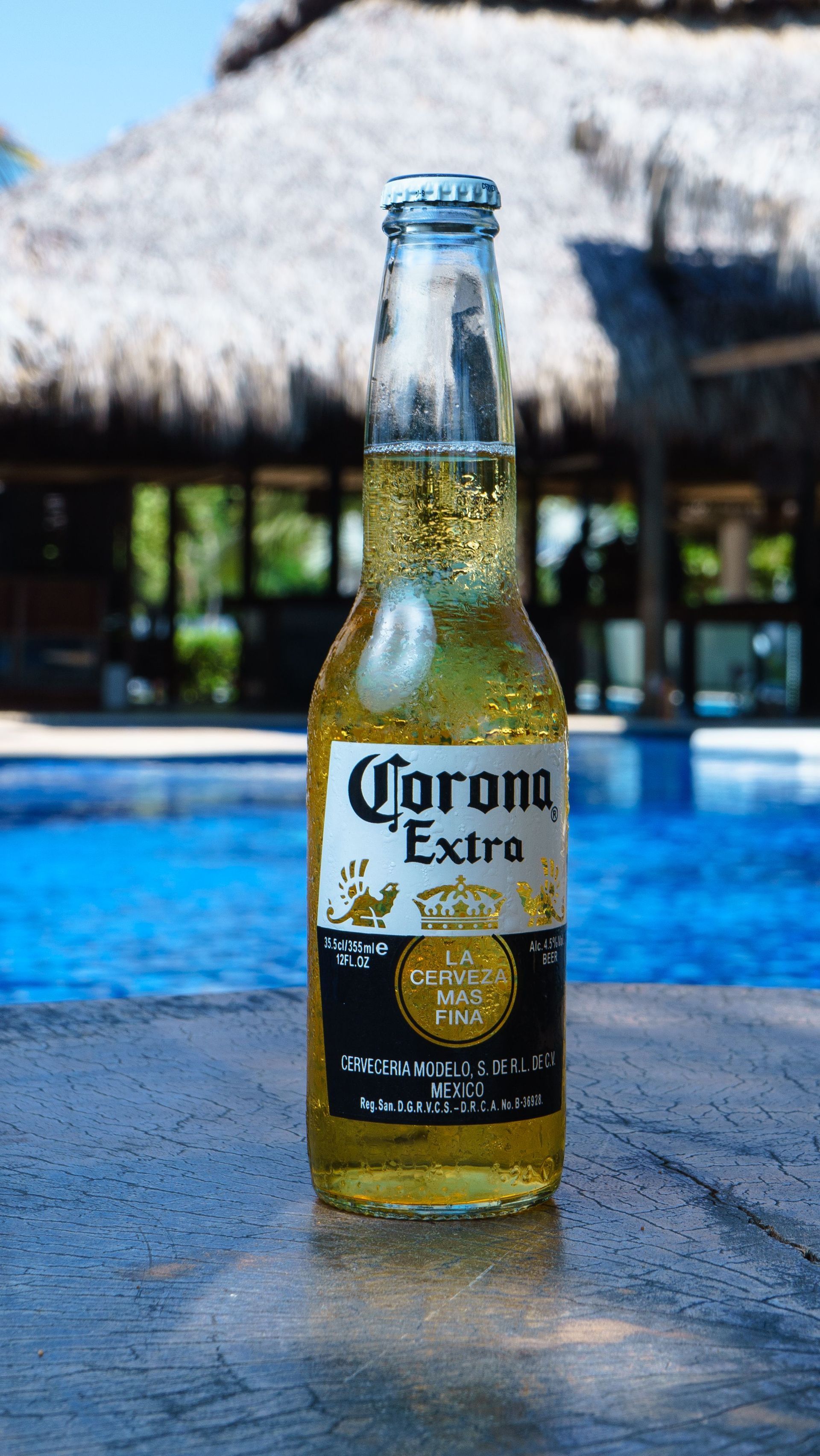 Corona Extra bottle on a table by a blue pool, with a thatched roof and outdoor bar in the background