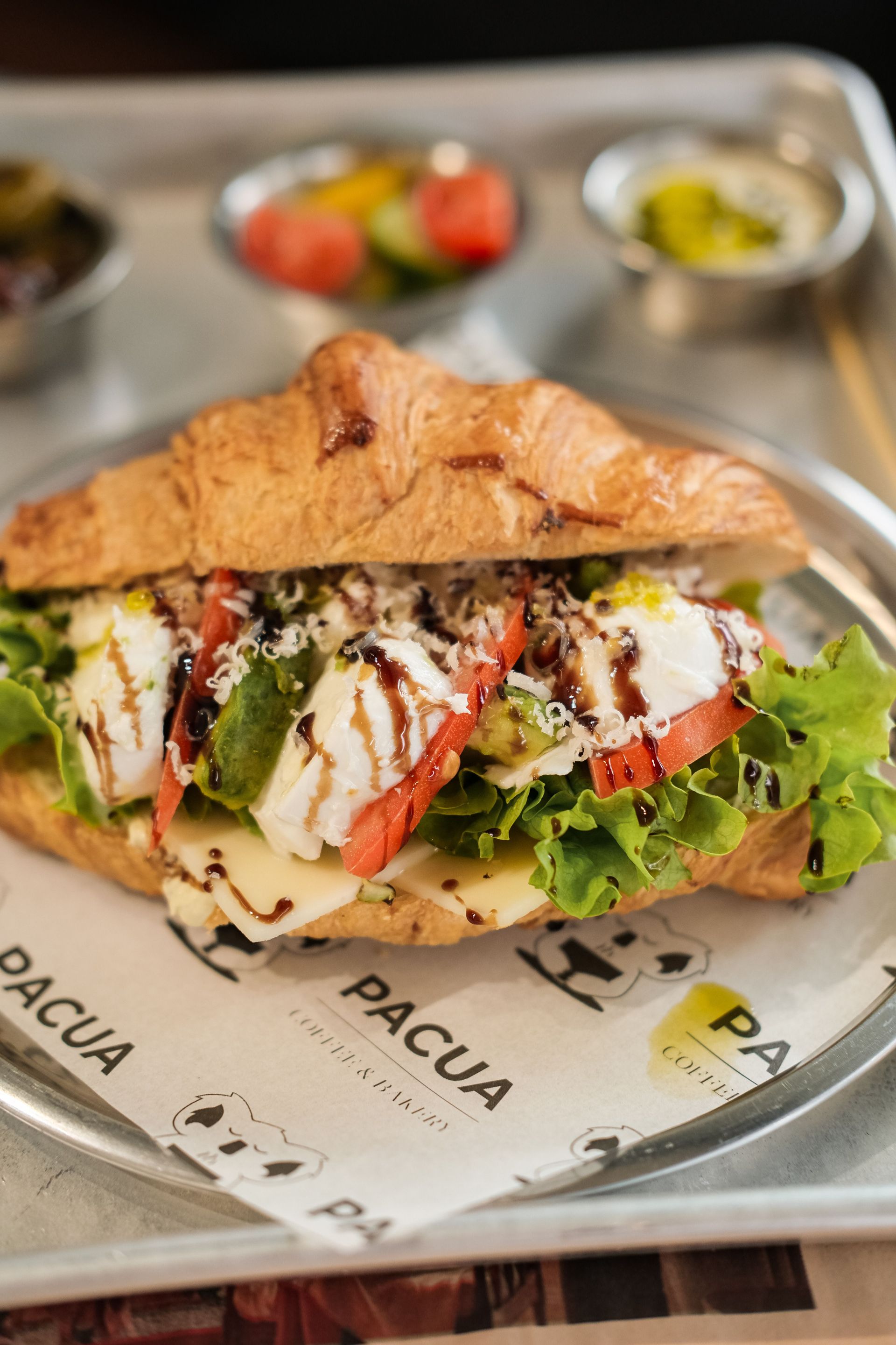 Grilled sandwich on a tray with greens, tomatoes, and drizzled sauce in a café setting