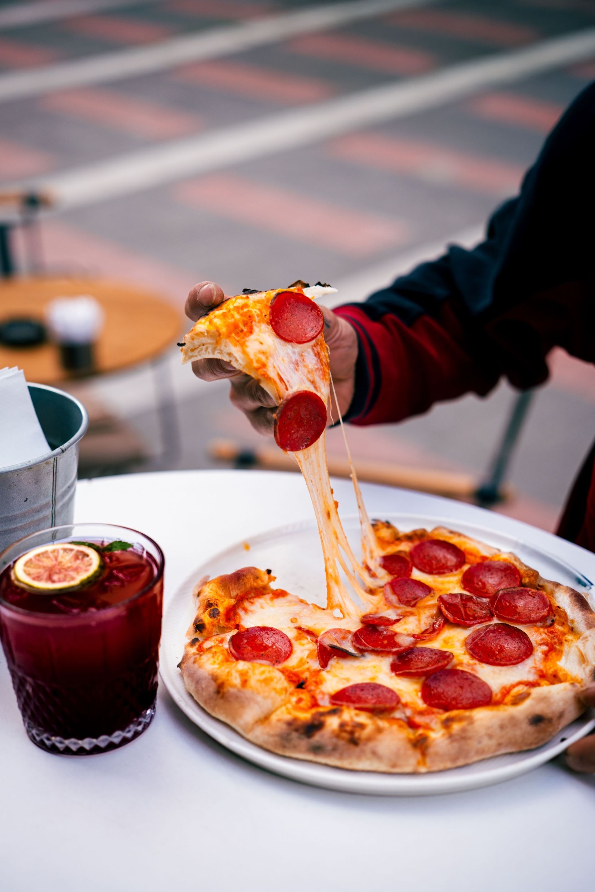 Hand holding a pepperoni pizza slice over a plate beside a drink on an outdoor café table
