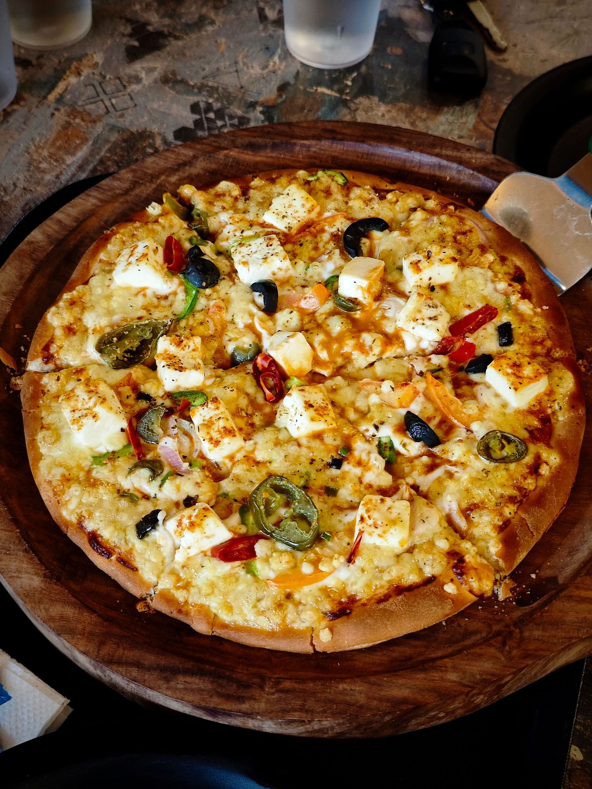 Pizza on a wooden board with melted cheese, vegetables, and red peppers at a restaurant table
