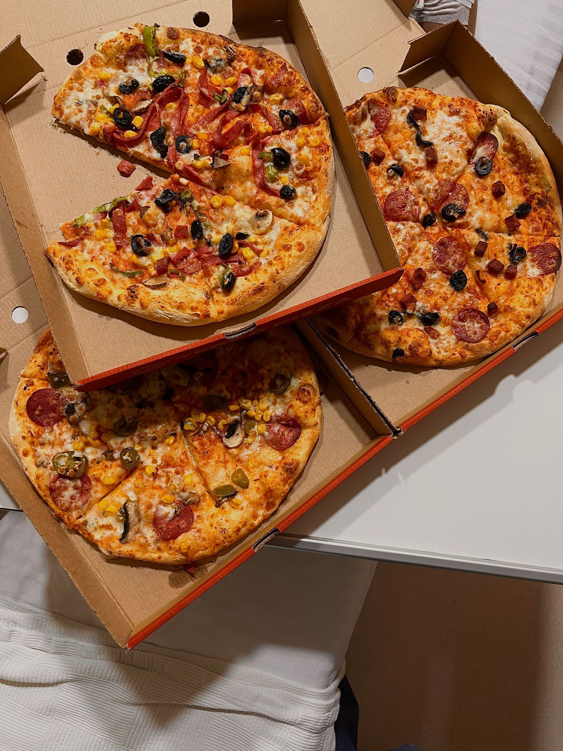 Three pizzas in open boxes on a table, topped with cheese, olives, and vegetables