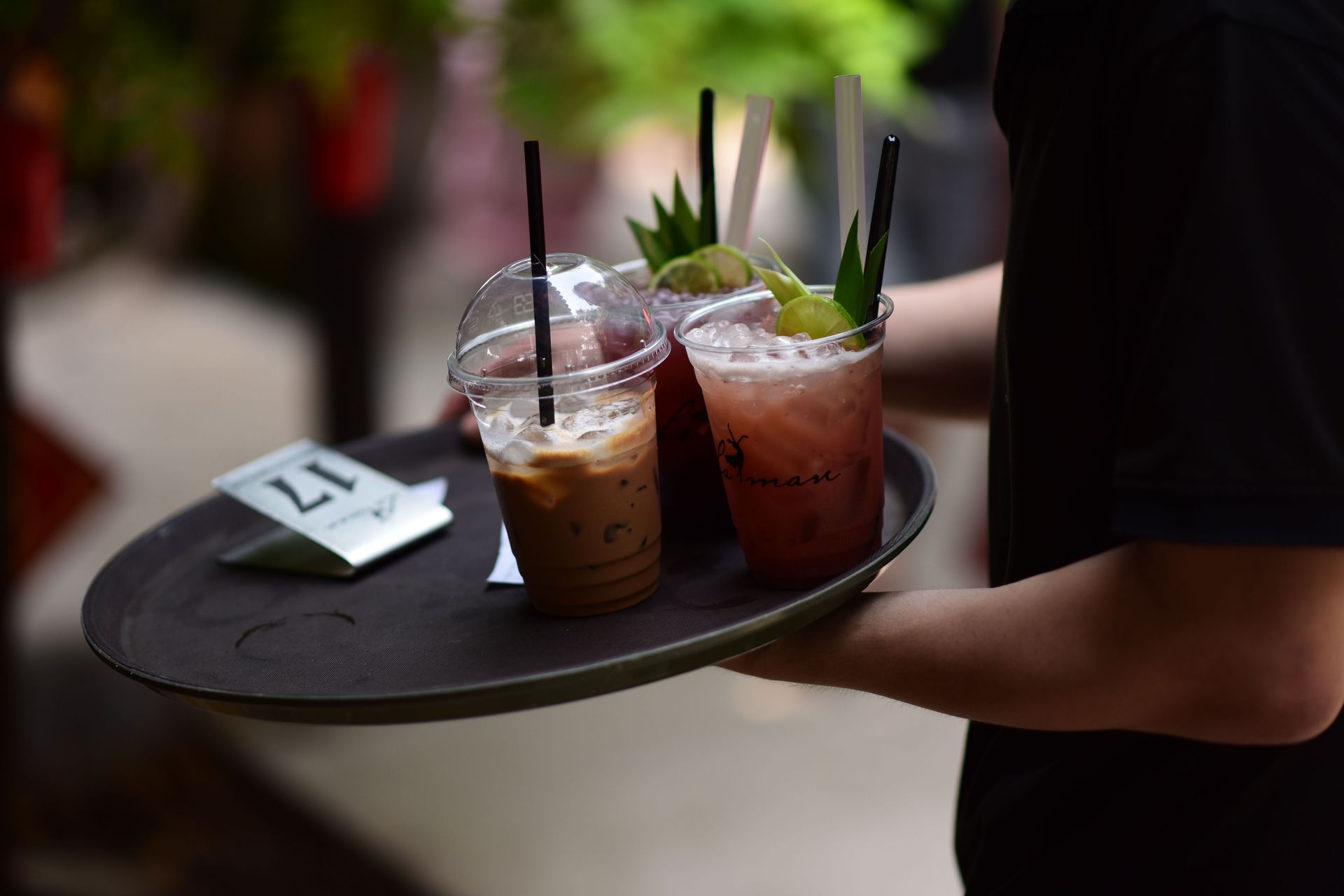 Hand holding a tray with two iced coffee drinks and straws, one topped with mint, in a café setting