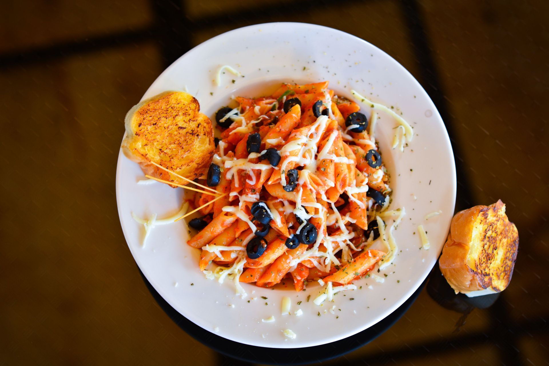 Top-down plate of pasta with orange sauce and black olives, with bread slices on a dark table.