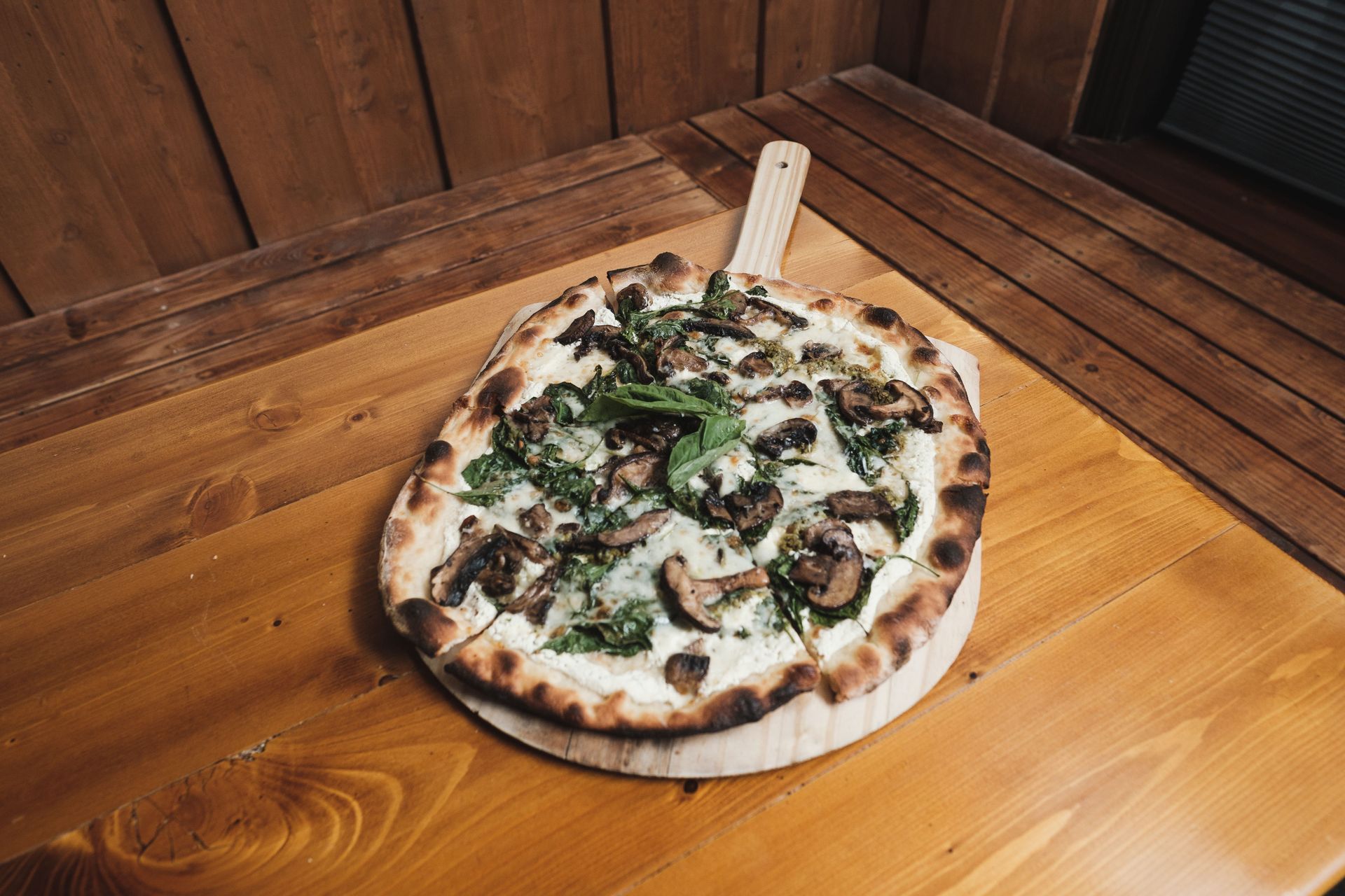 Pizza on a wooden board with greens and sliced toppings, set on a wooden table.