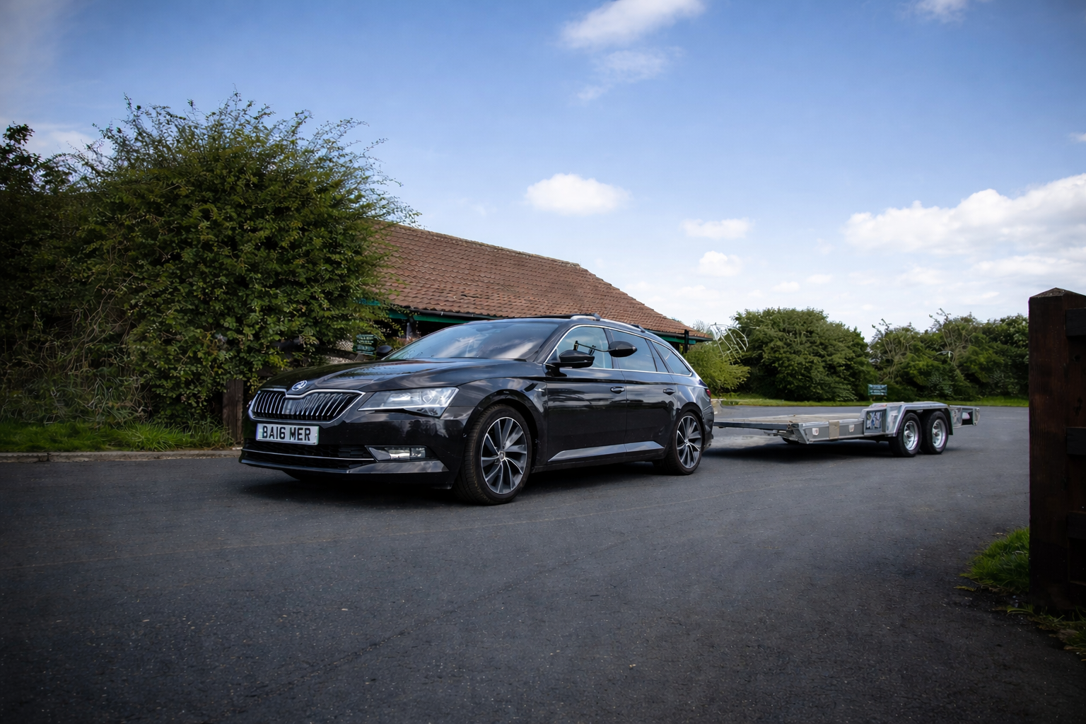 Black car towing a small trailer on a paved road, in front of a building with a red roof.