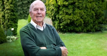 capecitabina precio Un anciano con un suéter verde está de pie con los brazos cruzados en un jardín.