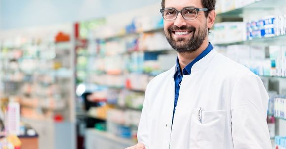 atendiendo pedidos de medicamento oncologico Farmacéutico sonriente con bata blanca de pie en un pasillo de la farmacia con estantes borrosos detrás de él.