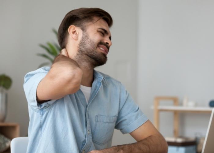 Un hombre con una camisa azul claro se frota el cuello mientras está sentado en el interior y sonríe.