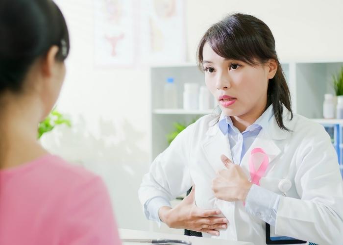 Un médico, vestido con bata blanca y lazo rosa, habla con un paciente en una clínica.