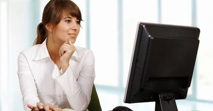 Mujer con camisa blanca mirando pensativamente un monitor de escritorio en una oficina luminosa.