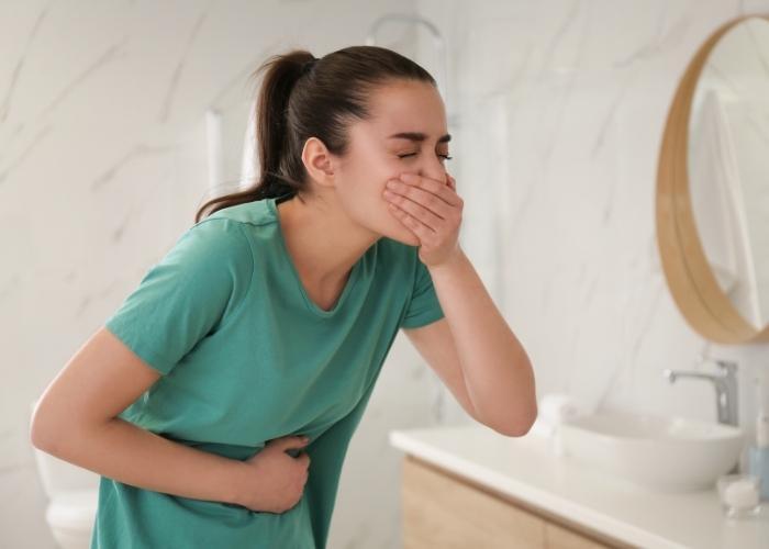 Mujer con camisa verde sujetándose el estómago y tapándose la boca en un baño, con aspecto de tener náuseas.