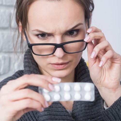 que efectos secundarios Mujer con gafas examinando un blíster de pastillas con expresión concentrada.