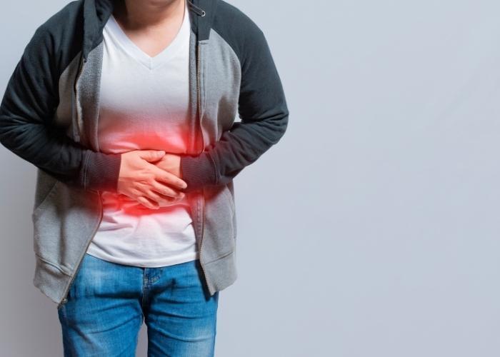 Persona que se sujeta la parte inferior del abdomen mostrando un signo de dolor intenso, vestida con un cárdigan gris y pantalones vaqueros azules.