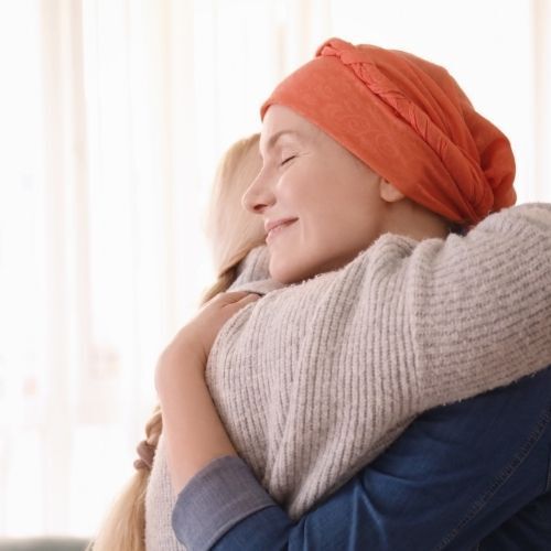 oncologia Dos personas se abrazan en el interior; una de ellas lleva un pañuelo naranja en la cabeza y sonríe con los ojos cerrados.