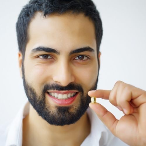 medicamento precio Hombre sonriente sosteniendo una pequeña píldora de oro entre sus dedos sobre un fondo blanco.