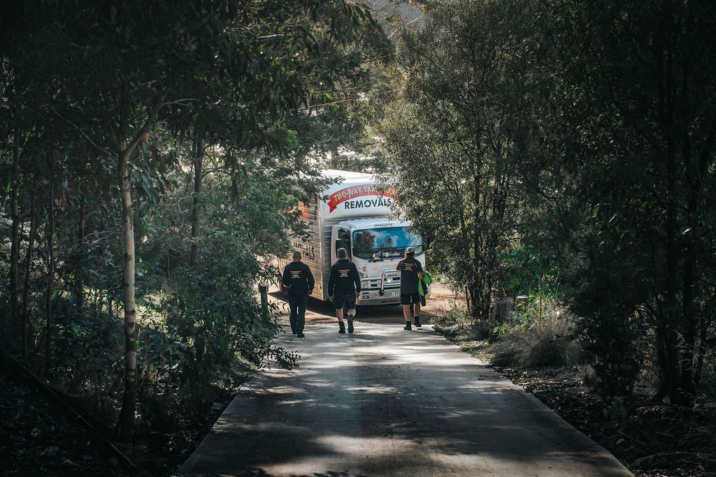 People walking on a tree-lined path toward a blue bus in a shaded park area