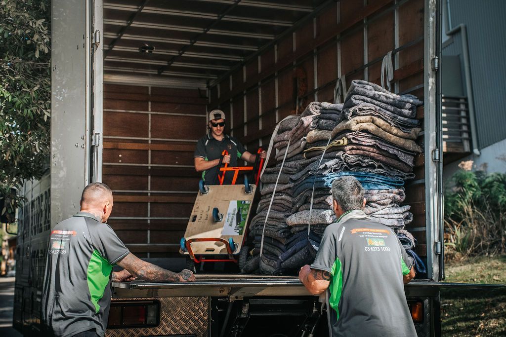 Three movers in grey and green uniforms load items and stacks of wrapped furniture into the back of a moving truck.