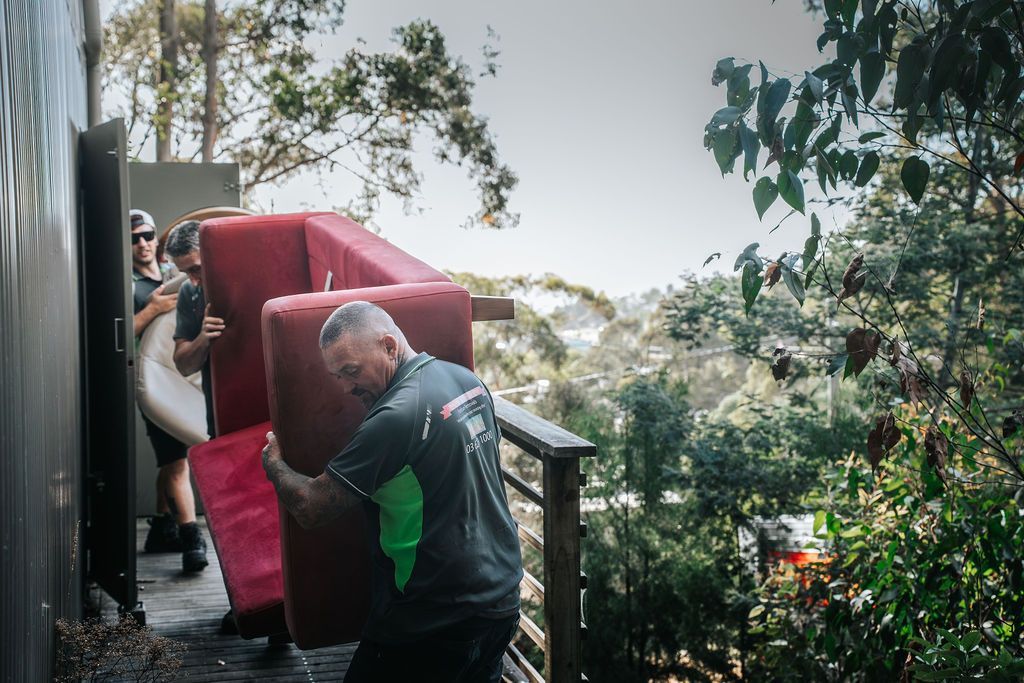 Two male movers lift a red couch while standing on a wooden deck.