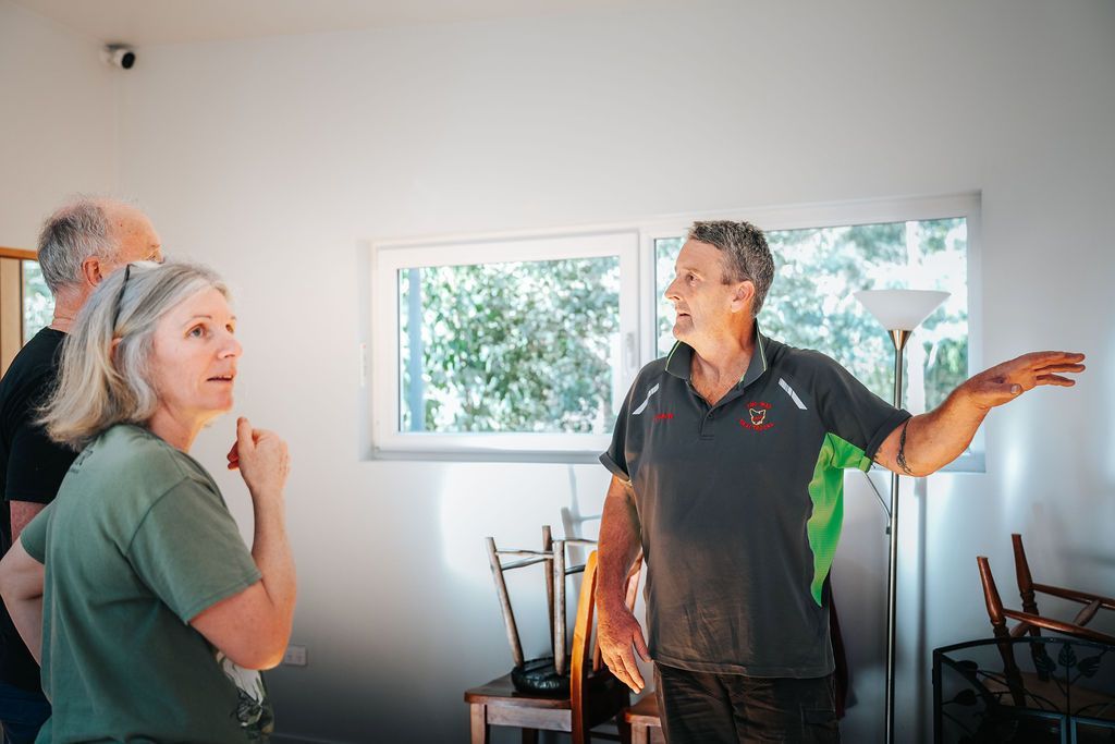 A person in a gray shirt gestures toward a window while talking to two other people in a bright, sparsely furnished room.