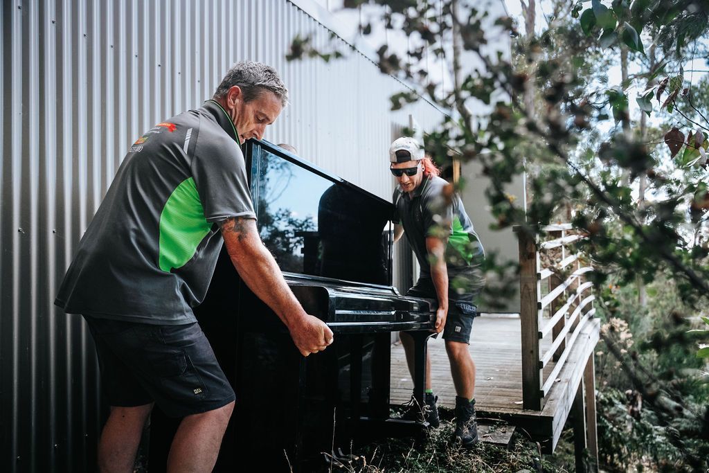 Two people moving a black piano outside beside a white building and a porch railing