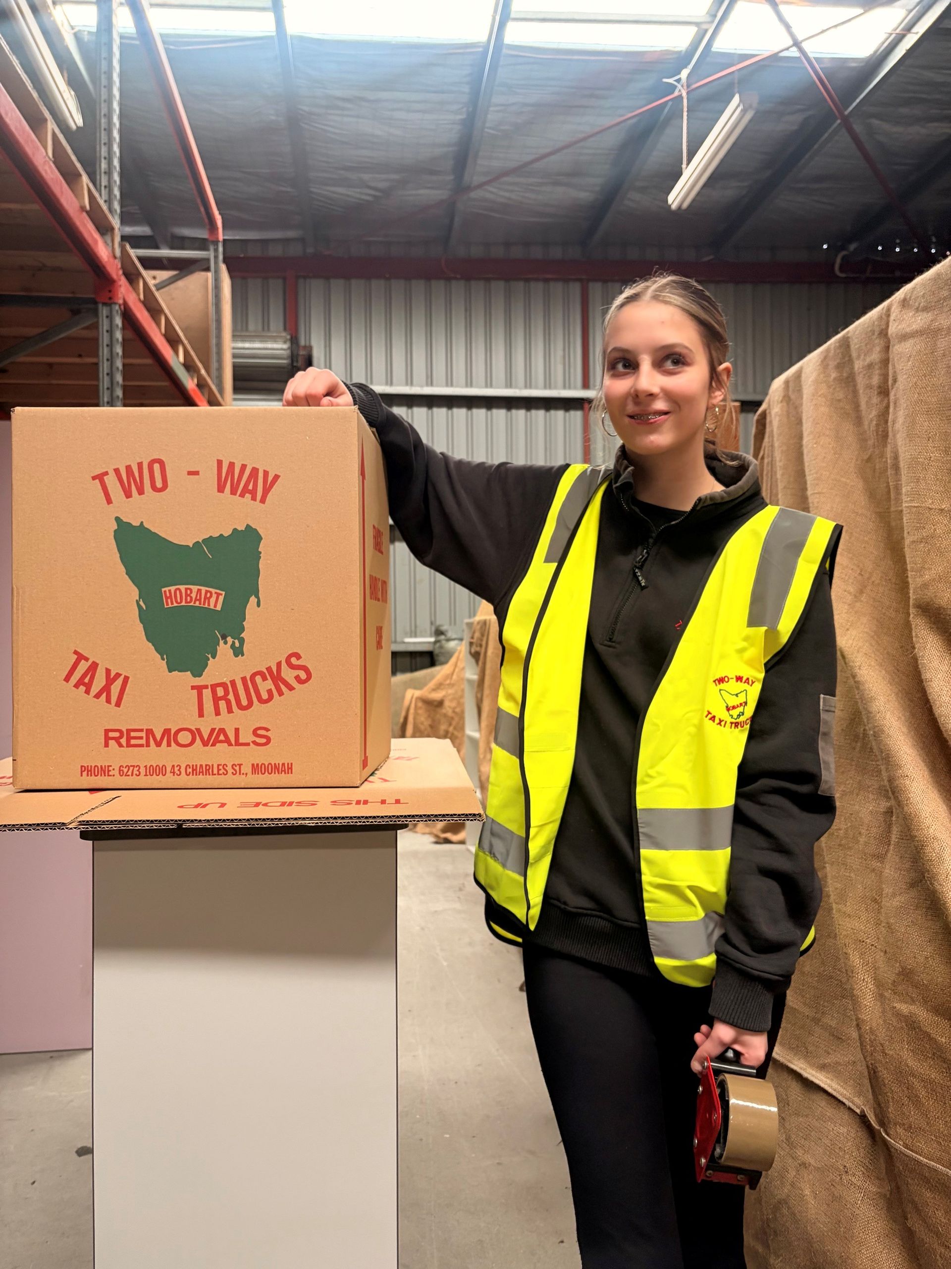 Woman in yellow safety vest beside a cardboard box labeled “All Star Trucks Removals” in a warehouse