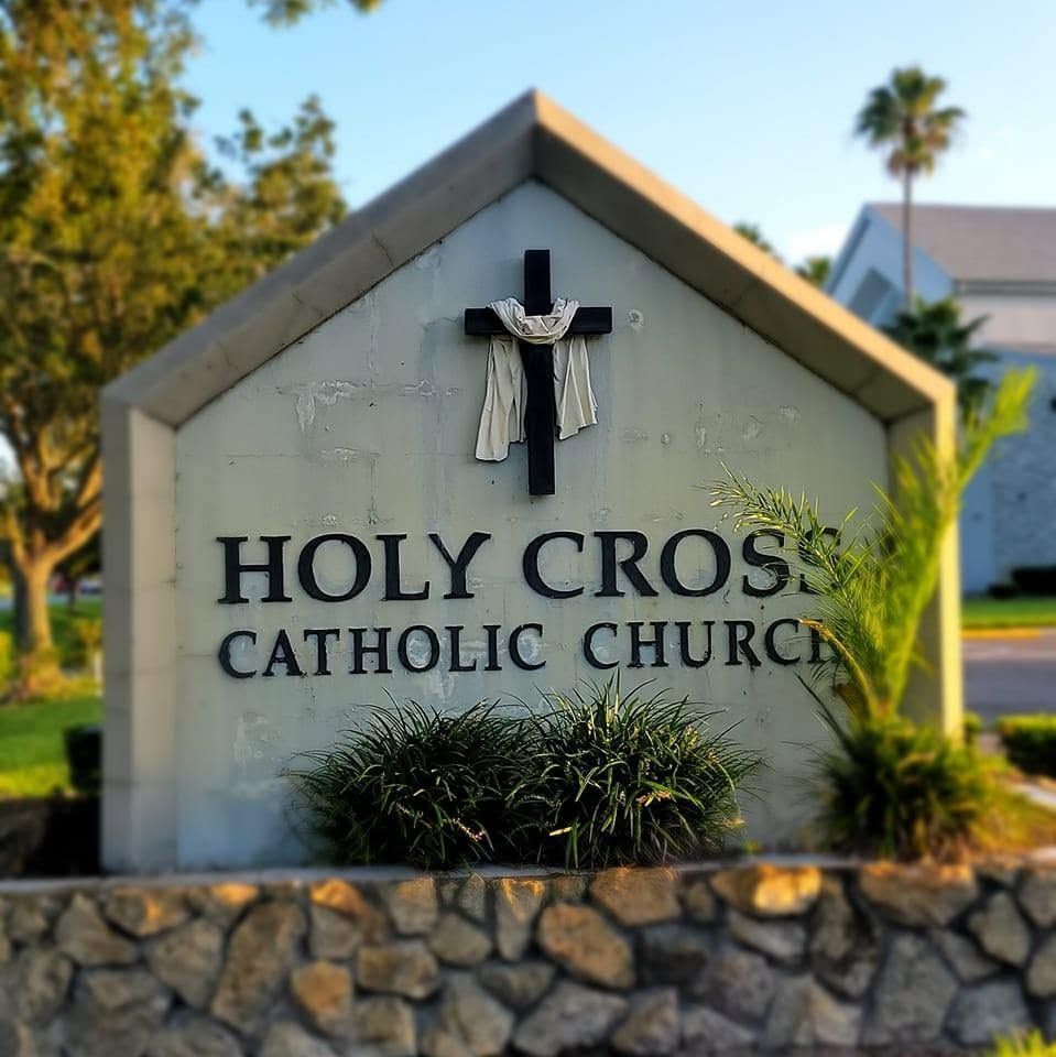 Holy Cross Catholic Church sign with crucifix on a stone wall, set among greenery