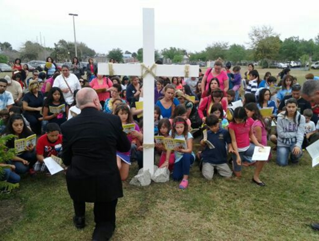 Large group seated outdoors around a white cross, with a person speaking to them in a grassy field.