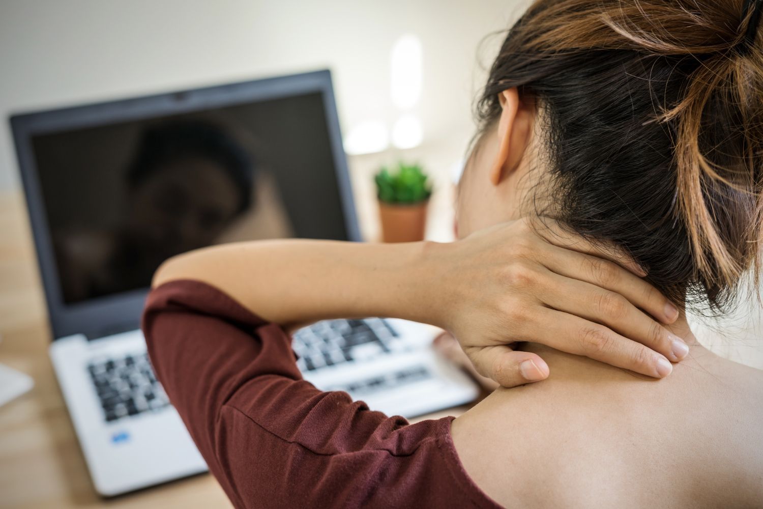 A woman is holding her neck in pain while sitting in front of a laptop computer.