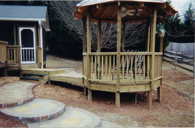 A wooden gazebo is sitting in the middle of a yard next to a house.