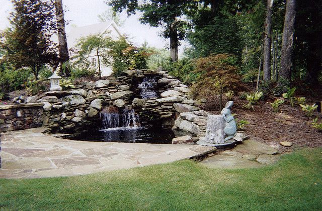 Garden waterfall with a stone pond, patio, and small statues surrounded by trees and grass