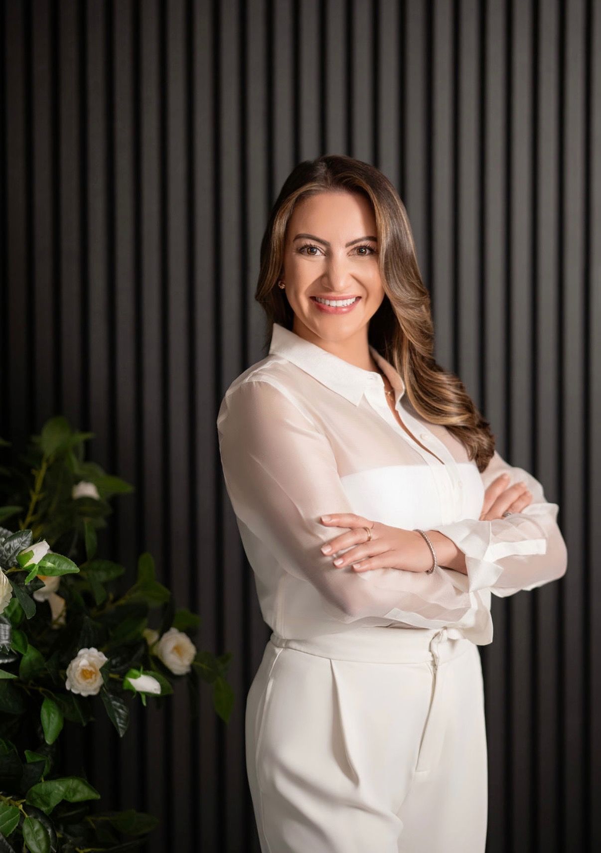 Woman in white outfit with arms crossed smiling in front of a gray textured wall, next to flowers.