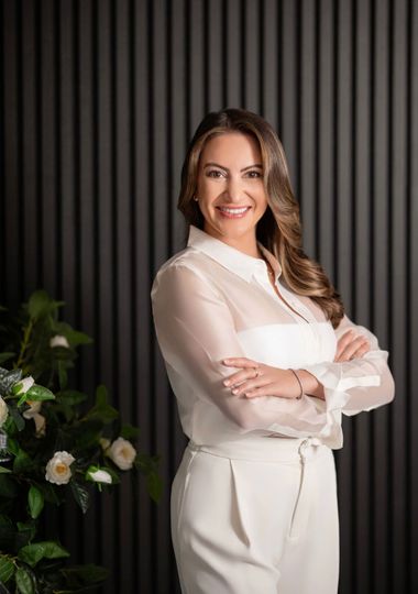 Woman in white outfit with arms crossed smiling in front of a gray textured wall, next to flowers.